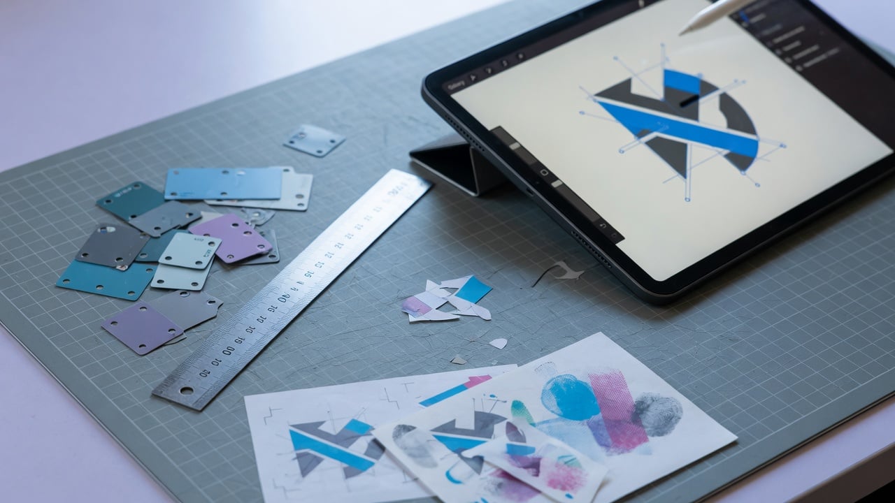 Designer at a slate desk with colour chips, a printed mark sketch, and a tablet showing abstract icon shapes